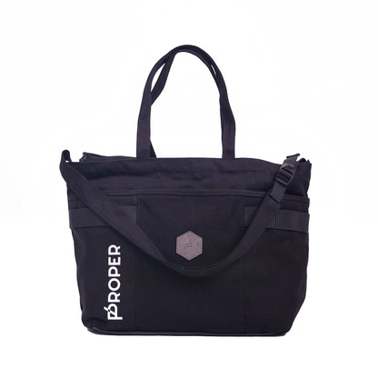 A black tote bag with the text 'PROPER' on the side, featuring a large main compartment, a front pocket, and adjustable straps.