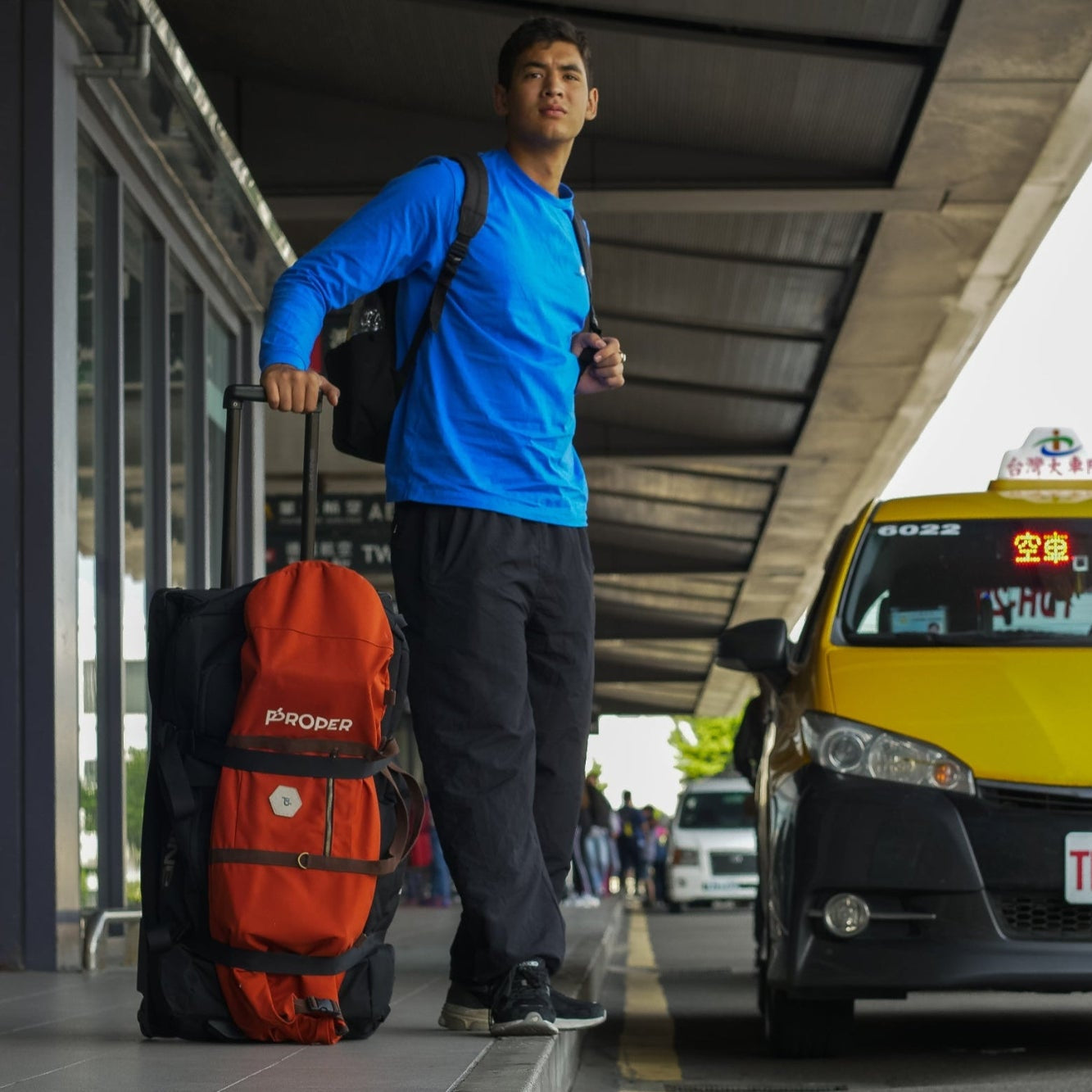 Man with a skateboard bag waiting at a taxi stand with a yellow taxi in the background at the airport