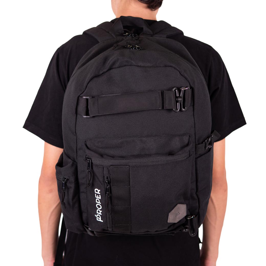 A person wearing a black T-shirt and black shorts is seen from the back, carrying a large black canvas backpack with multiple compartments and secure straps.