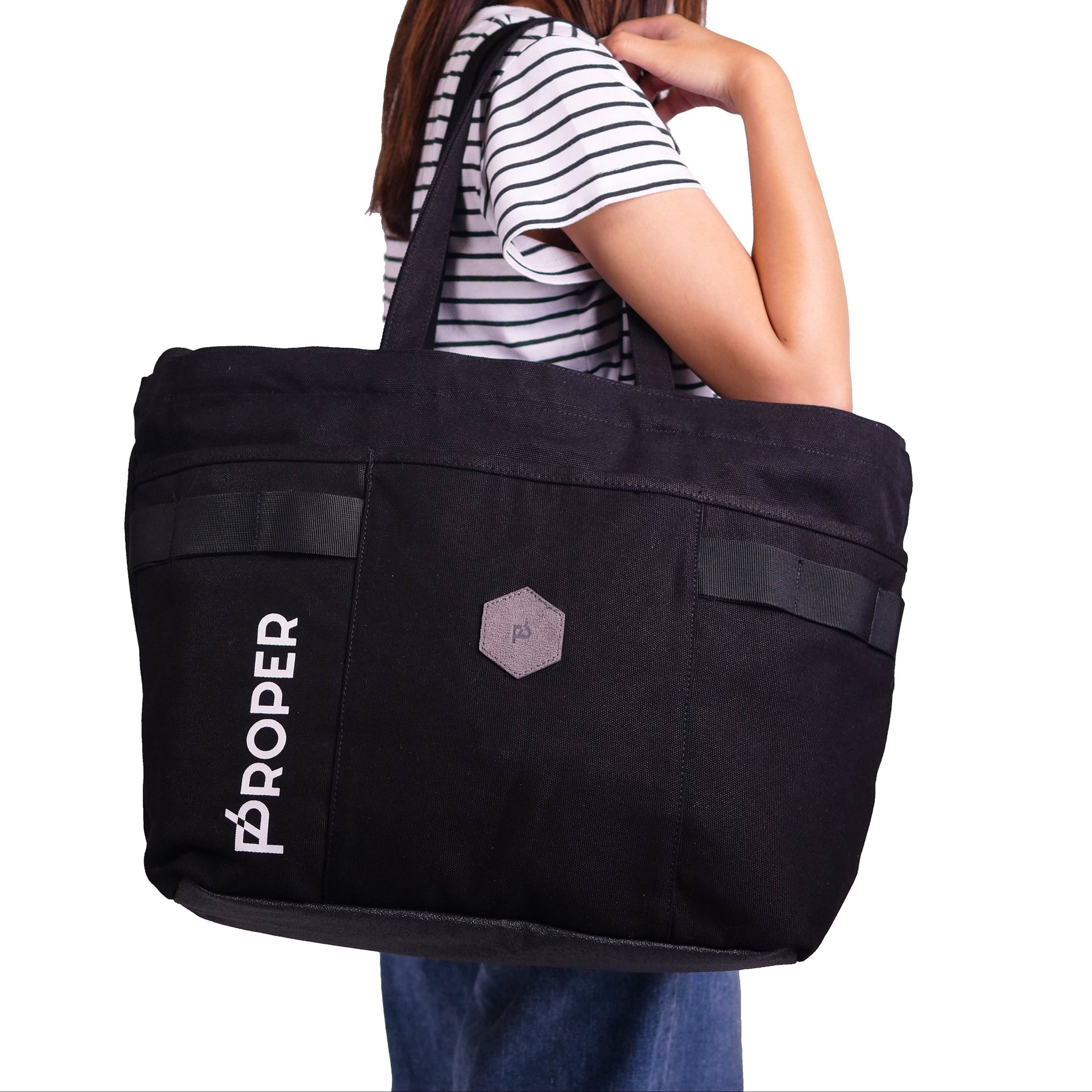 A black tote bag with the text 'PROPER' on the side, featuring a large main compartment, a front pocket, and adjustable straps.