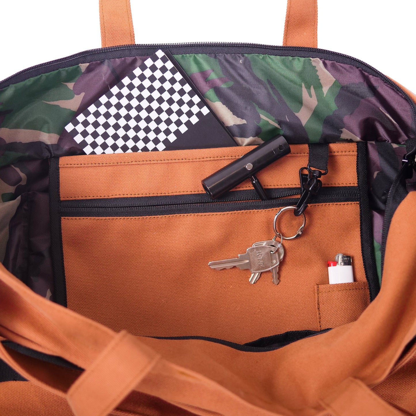 Close-up of the inside of a tote bag showing a brown front pocket with zipper, a black flashlight, keys on a key ring, and a small white item in a side pocket. The bag interior has a green and purple camouflage pattern and orange-brown handles.
