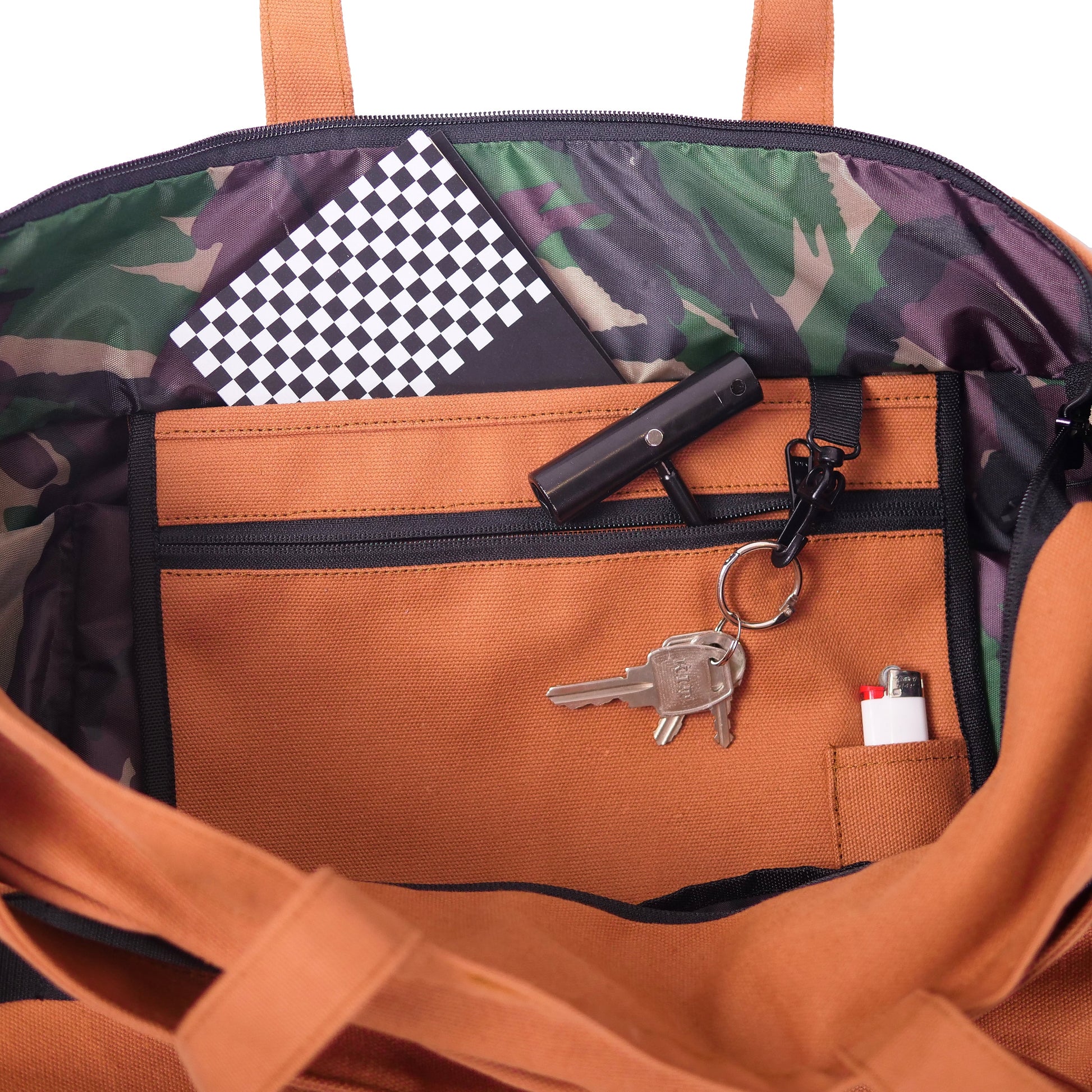 Close-up of the inside of a tote bag showing a brown front pocket with zipper, a black flashlight, keys on a key ring, and a small white item in a side pocket. The bag interior has a green and purple camouflage pattern and orange-brown handles.